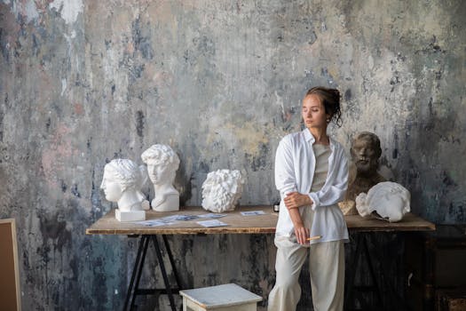 Jeune artiste en tenue blanche se tenant près d'une table avec des sculptures en marbre et des croquis sur un mur texturé.